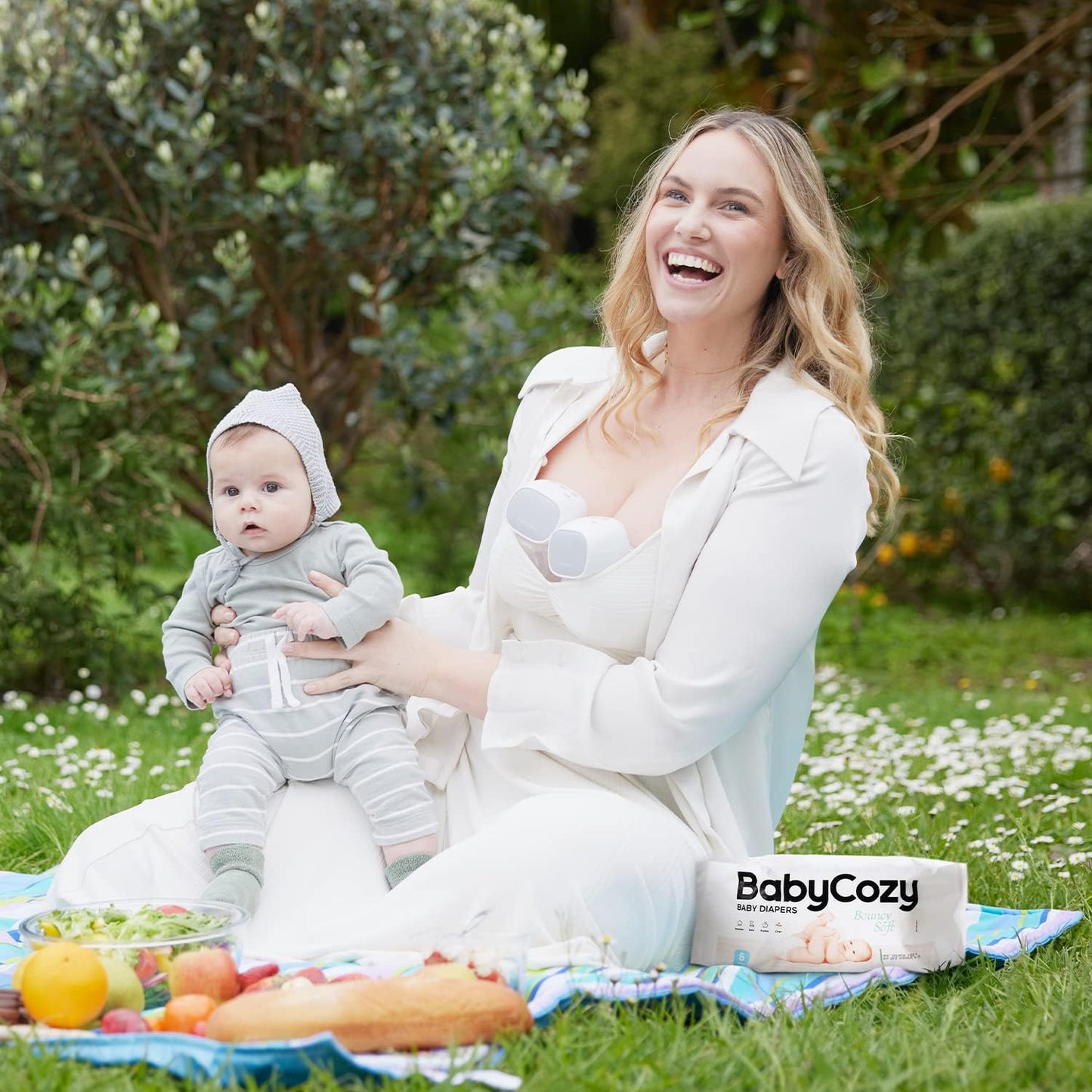 Mother holding baby outdoors wearing Babycozy size 3 diapers with a picnic spread on the grass