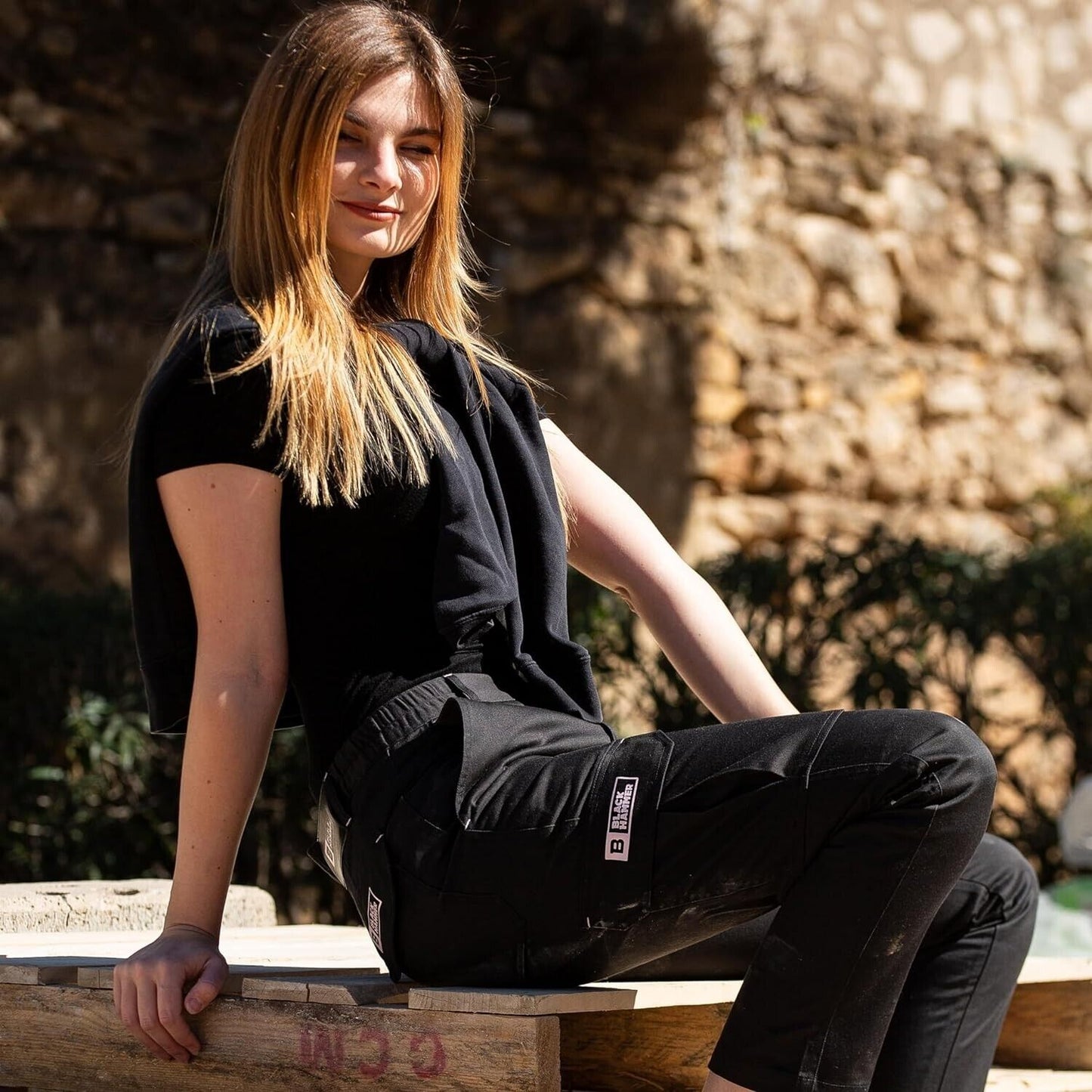Woman wearing black hammer cargo pants size 14 sitting outdoors with a stone wall background