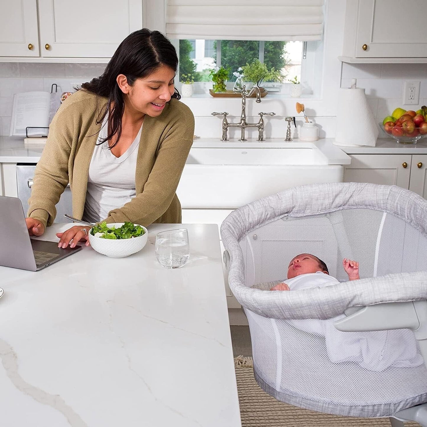 A woman working on a laptop at the kitchen table next to a halo baby bassinet sleeper with a sleeping baby