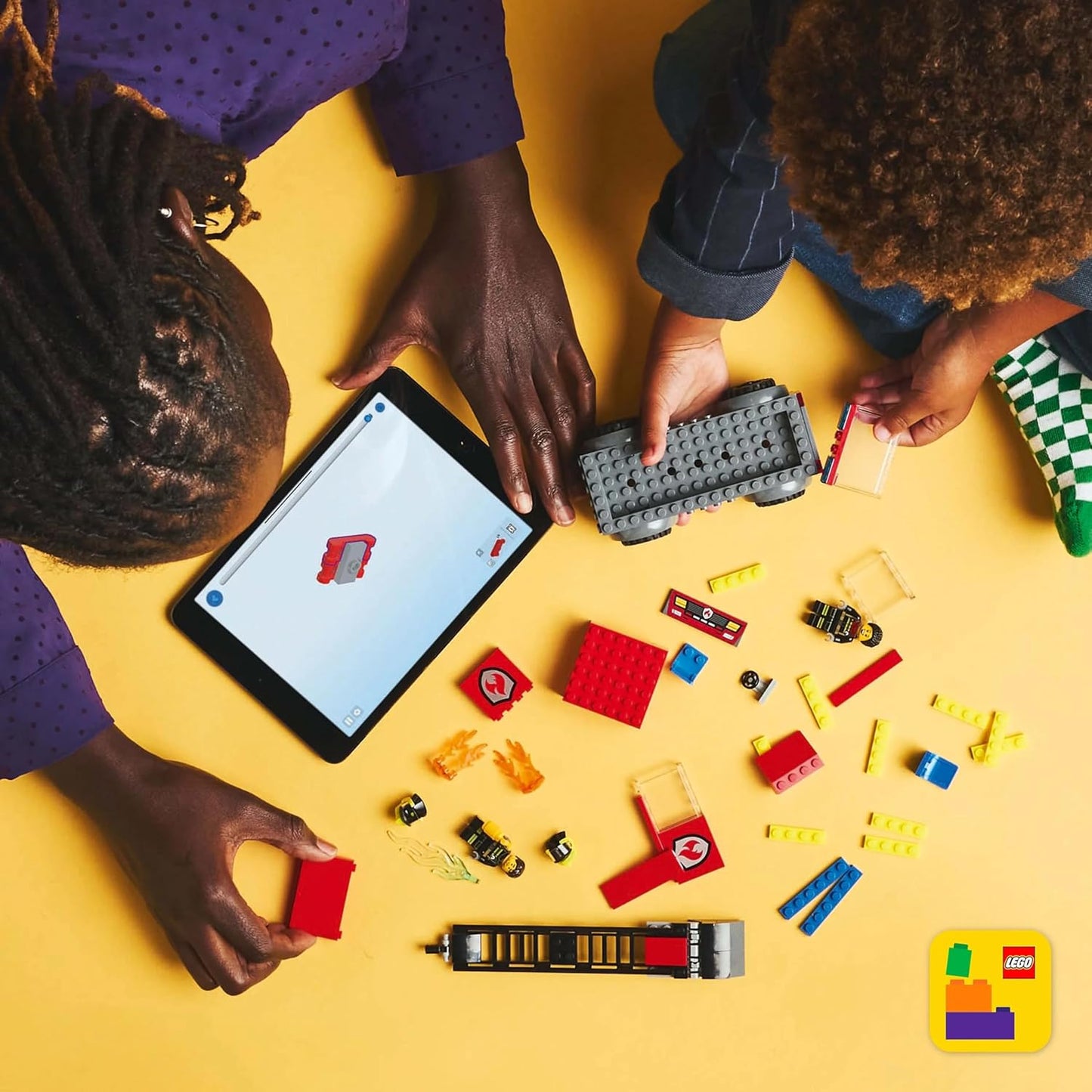 Children playing with LEGO City Fire Engine toy pieces and a tablet on a yellow surface