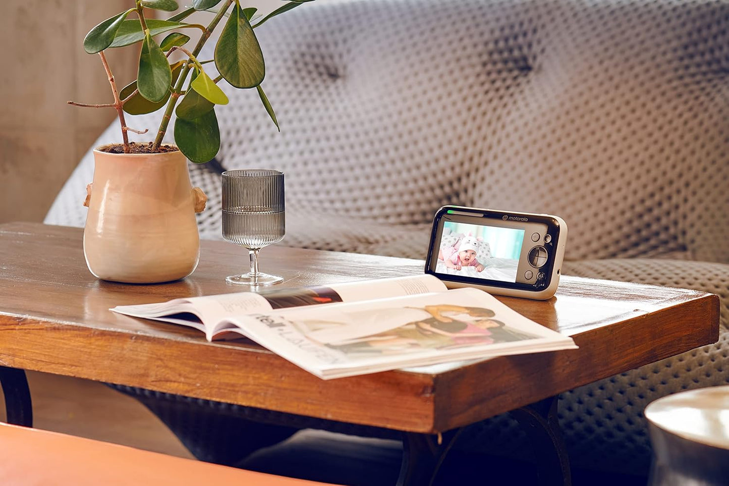 Motorola video baby monitor showing a baby on screen, placed on a stylish wooden table with a plant and glass.