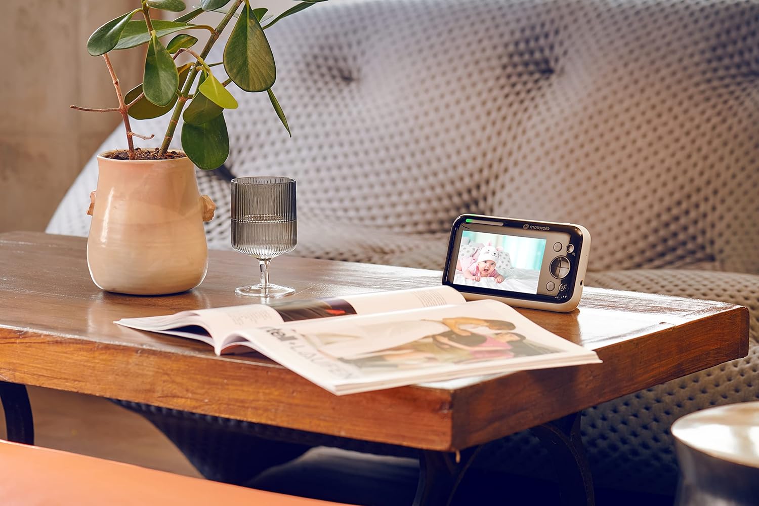 Motorola video baby monitor showing a baby on screen, placed on a stylish wooden table with a plant and glass.
