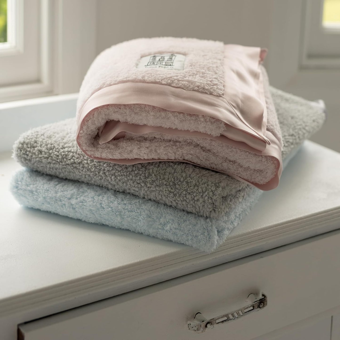 Soft blue baby chenille blanket stacked with pink and grey blankets on a white shelf