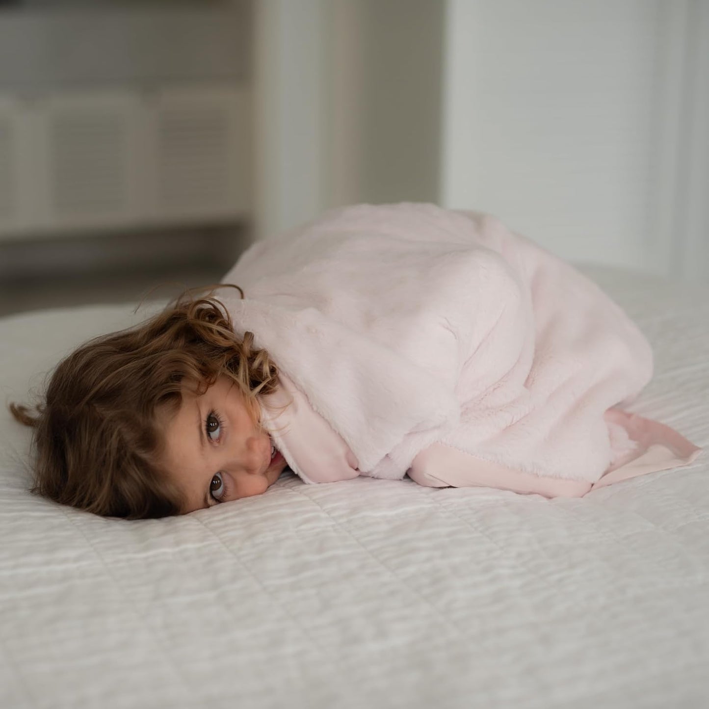 A child snuggled in a soft pink baby chenille blanket on a white bed