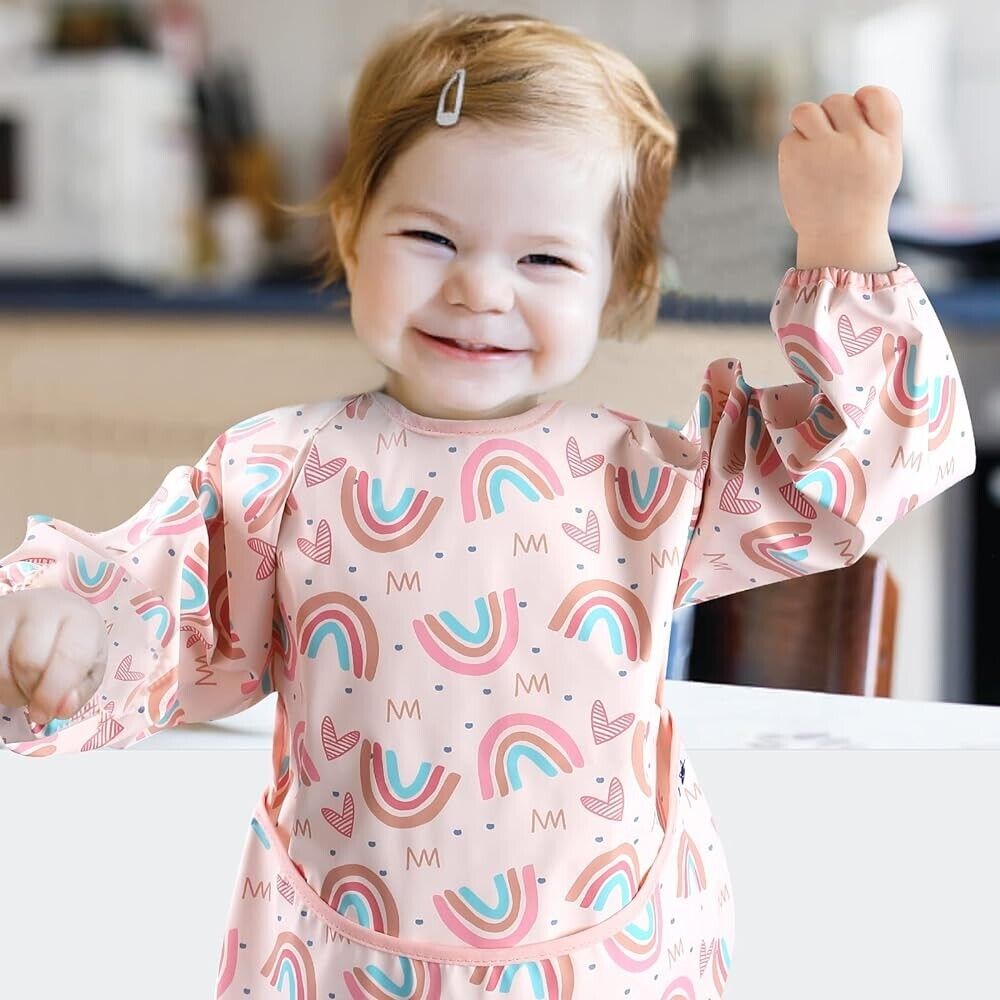 Cute toddler wearing long sleeve weaning bibs with colourful rainbow design in a bright kitchen