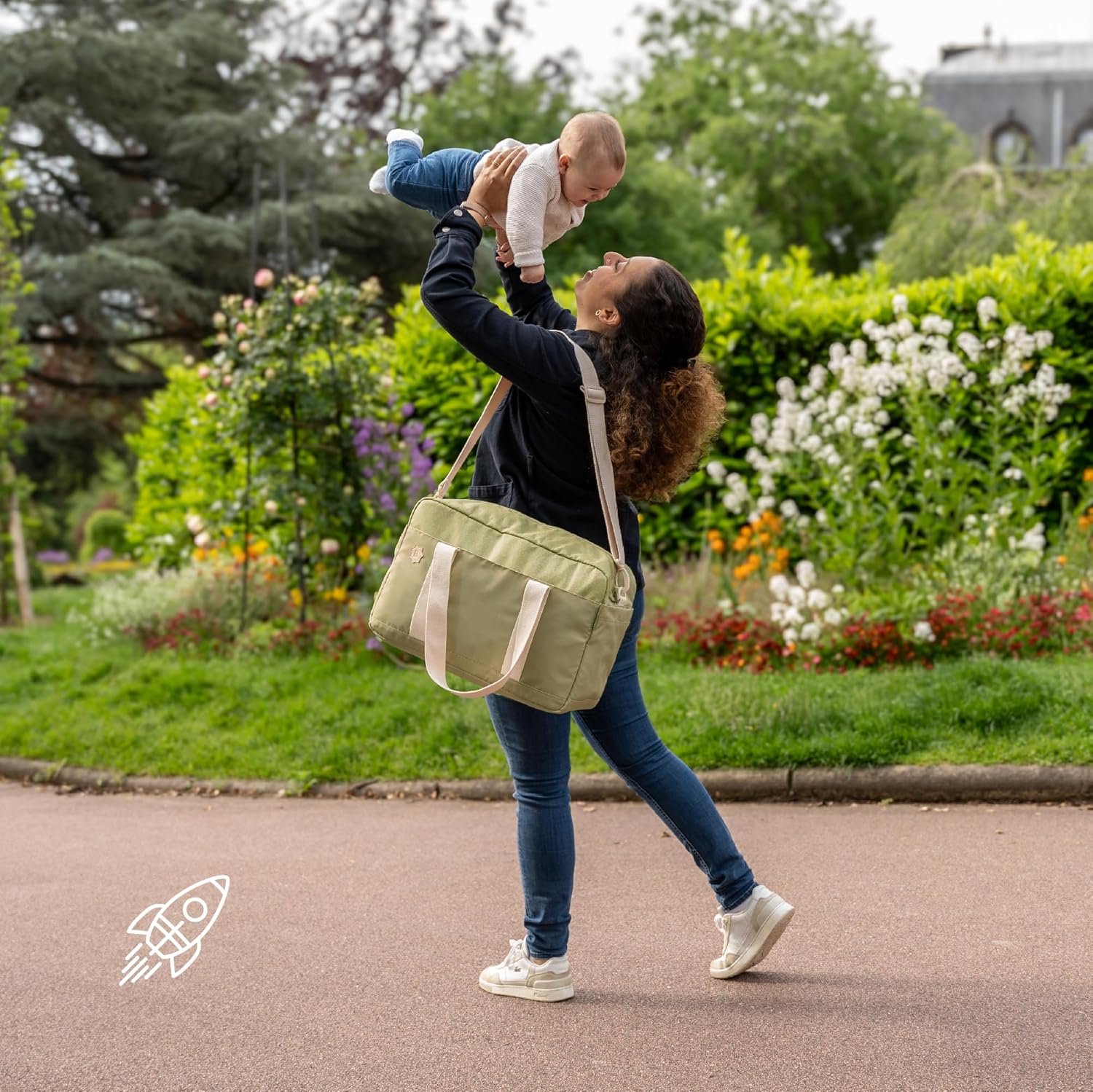 Mother holding a baby while wearing an olive baby changing bag in a park setting