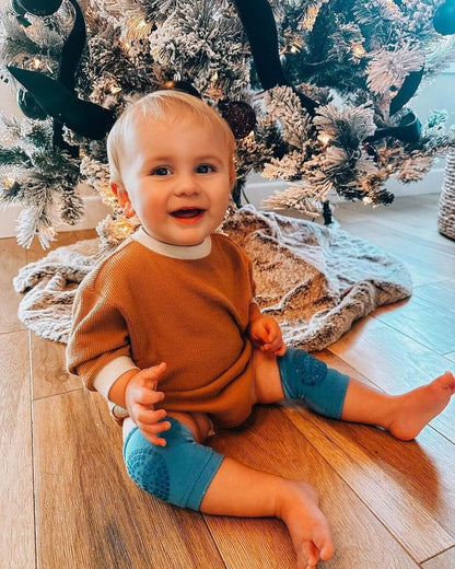 Happy baby in a cosy outfit wearing anti-slip baby knee pads and socks set while sitting by a Christmas tree